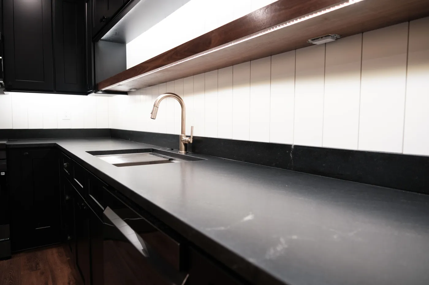 Black shaker cabinets with honed charcoal countertops, white vertical shiplap backsplash, walnut floating shelf, and a brushed brass pull-down faucet.