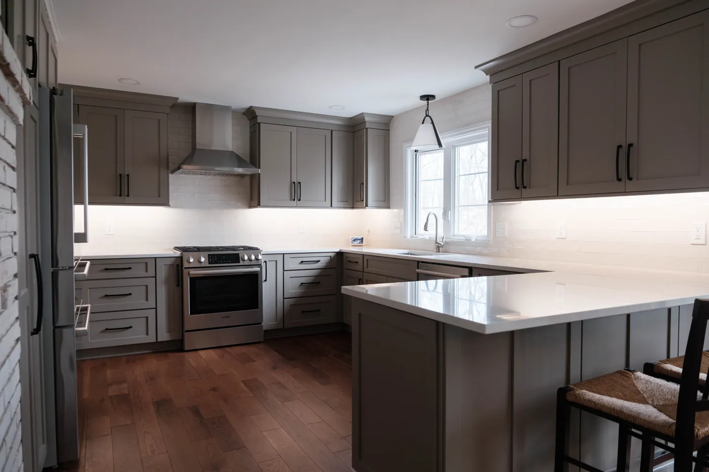 Gray shaker cabinets with black hardware, white subway backsplash, stainless range and hood, white quartz peninsula, and dark hardwood floors.