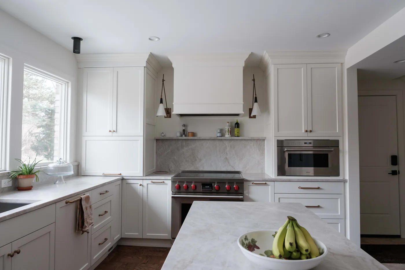 Creamy white cabinets, marble backsplash and counters, a red-knob pro range under a wood-framed hood, and brass sconce lighting.