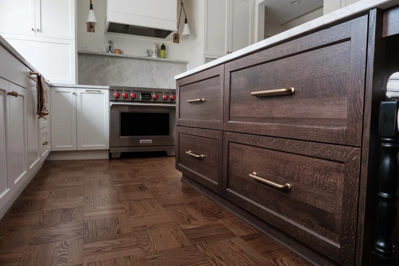Dark stained oak island with brass bar pulls in the foreground, herringbone wood floor, and a stainless range against white cabinetry.