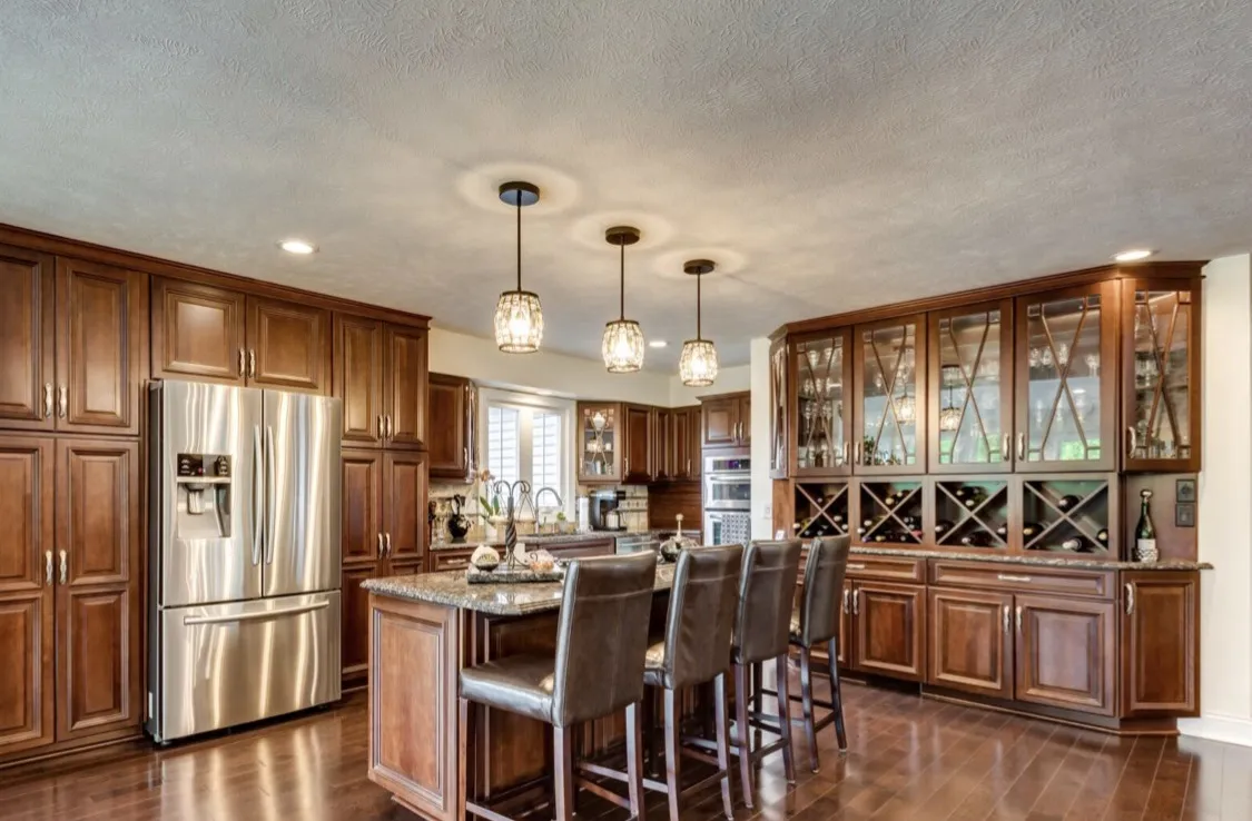 Traditional cherry-stained cabinets with glass-front wine storage, granite island seating four leather chairs, and a stainless French-door refrigerator.