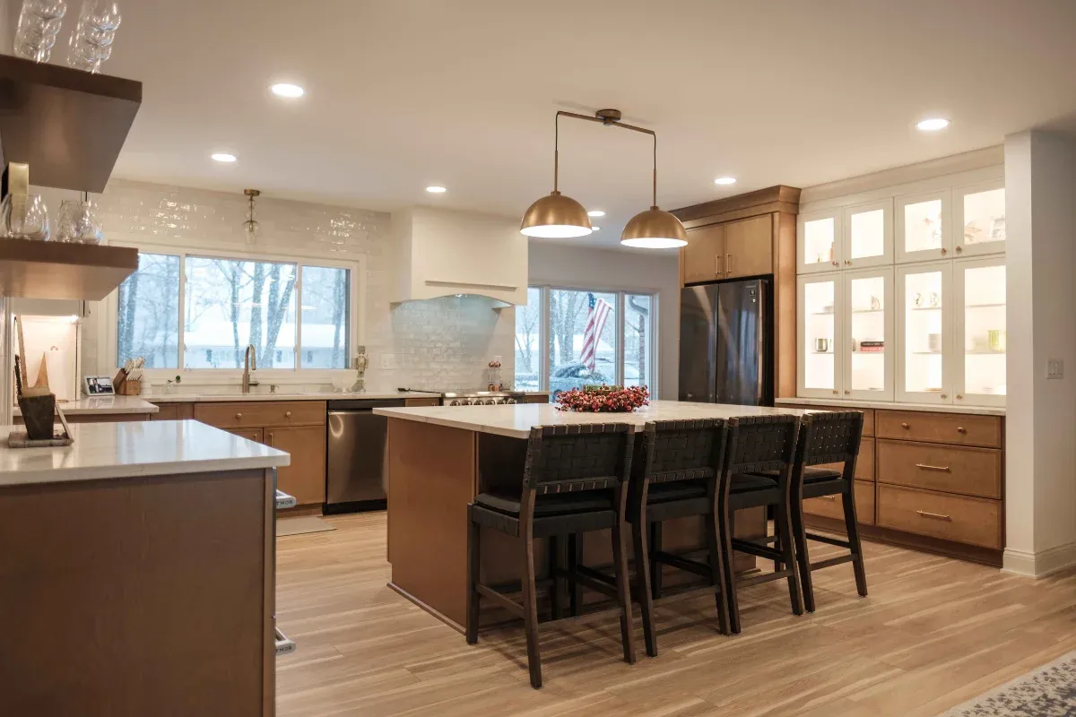 Two-tone kitchen mixing warm maple base cabinets and white glass-front uppers, brass dome pendants, and a white-topped island with woven stools.
