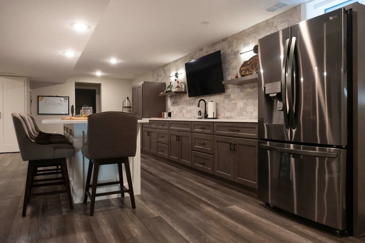 Basement bar kitchenette with gray-brown shaker cabinets, stone tile backsplash, mounted TV, black stainless refrigerator, and gray plank flooring.