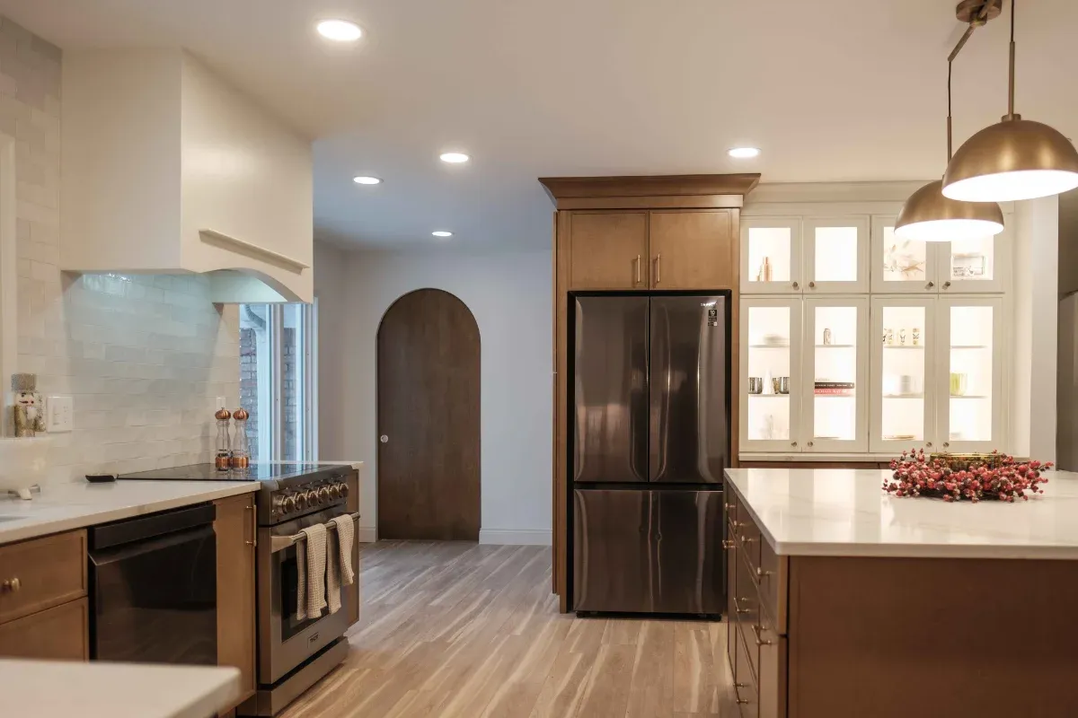 Wide view with medium-stained cabinets, waterfall-edge white island, arched wood door, lit white glass-front hutch, and brass dome pendants.