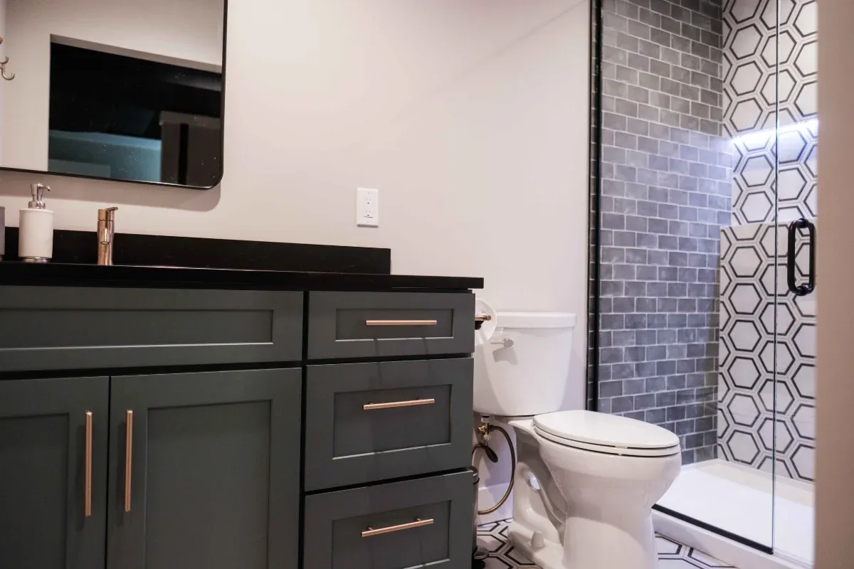 Dark green shaker vanity with a black countertop and brushed-gold pulls, beside a gray subway-tile shower with a hexagon accent wall.