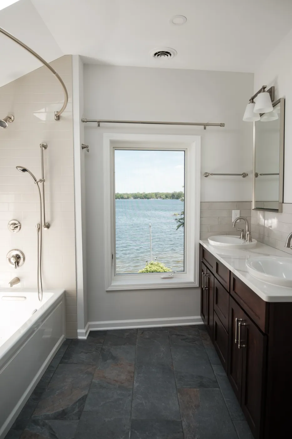 Lakeview bathroom with a white subway-tiled tub-shower, dark wood double vanity, slate floor tile, and a window overlooking open water.