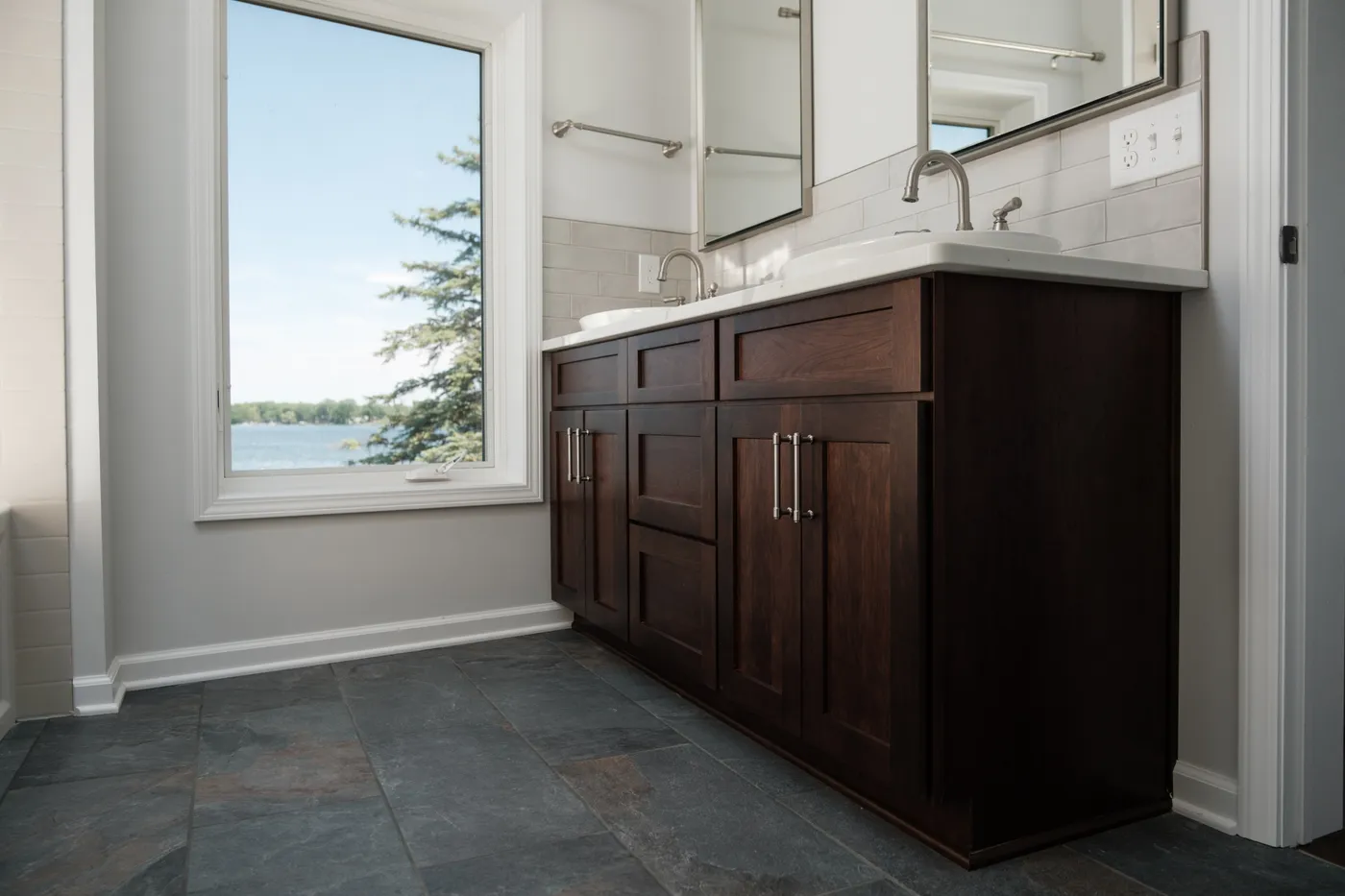 Espresso-stained double vanity with white countertop and brushed-nickel faucets against subway tile, slate floor, and a window framing a lake.