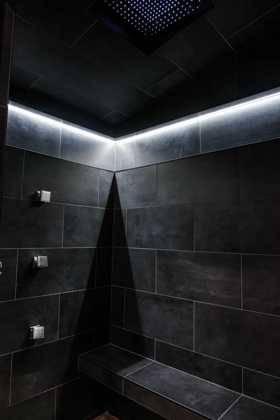 Dramatic black-tiled steam shower with LED cove lighting, a ceiling rain panel, chrome body-spray jets, and a built-in corner bench.
