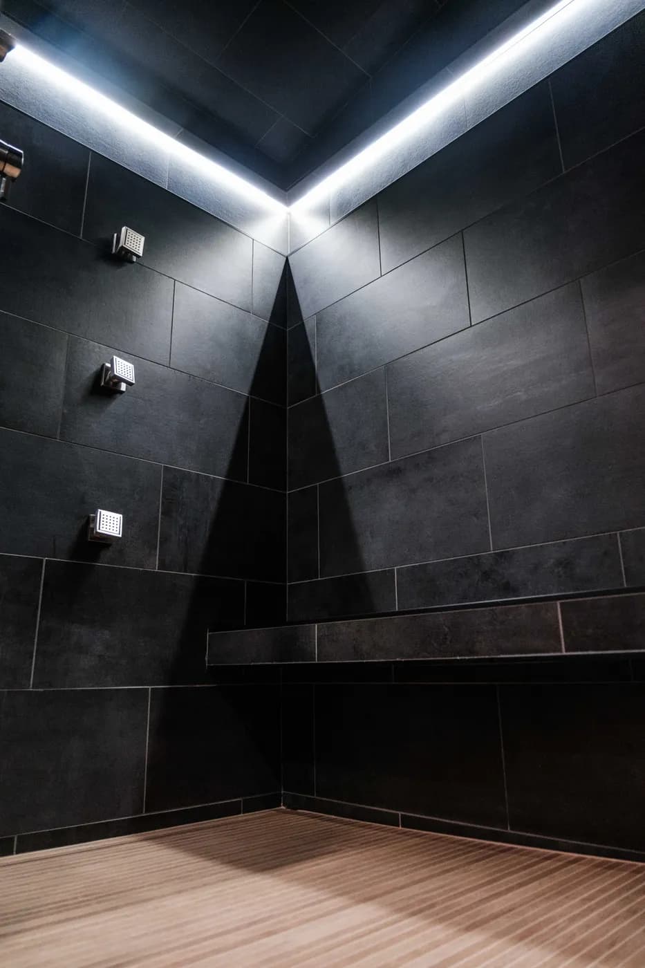 Corner of a charcoal-tiled shower with three chrome body sprays, glowing LED perimeter lighting, a floating tile bench, and wood-look plank floor.