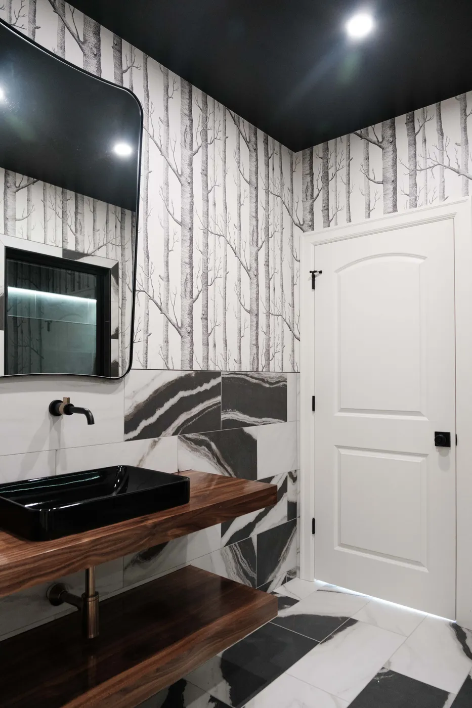 Bold powder room with birch-tree wallpaper, black ceiling, glossy black vessel sink on a walnut shelf, and black-and-white marble tile.