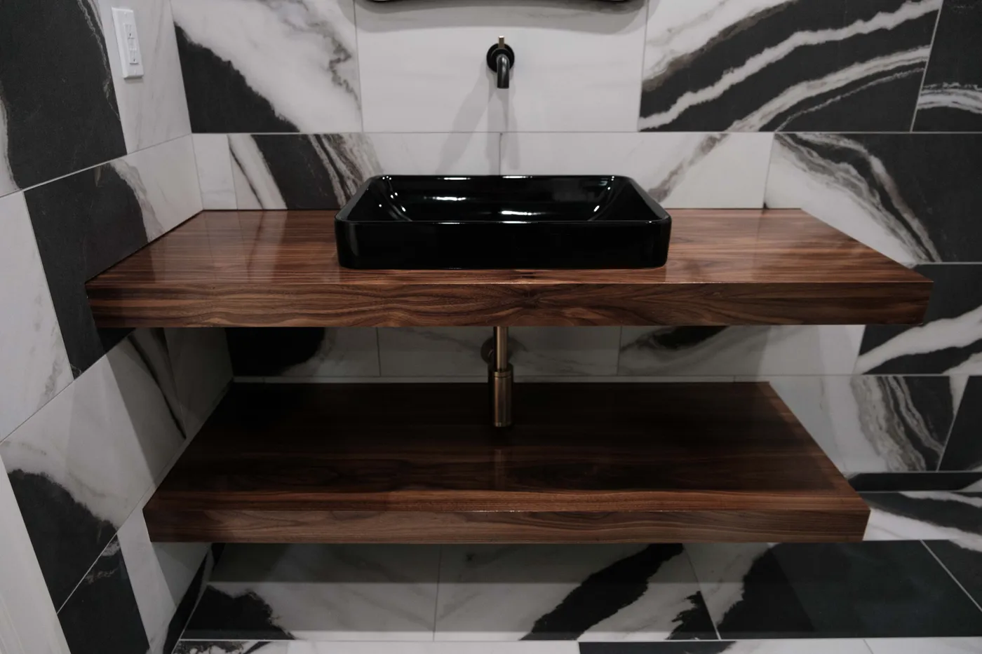 Close-up of a glossy black rectangular vessel sink on stacked walnut floating shelves, with brass plumbing against black-and-white marble tile.