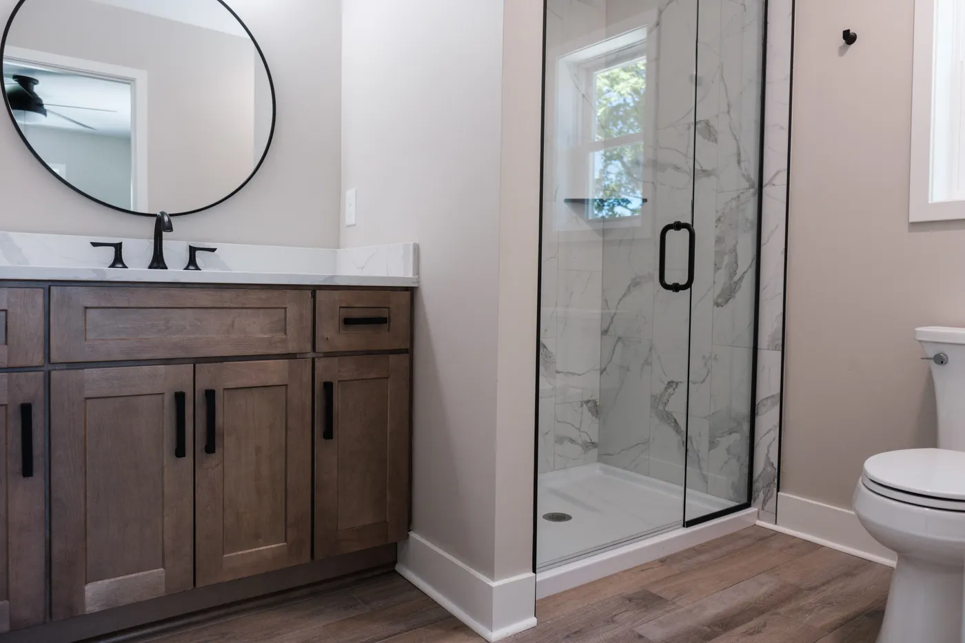 Bright bath with a stained-wood vanity, white marble countertop, round black-framed mirror, and a marble-tiled glass shower beside a white toilet.