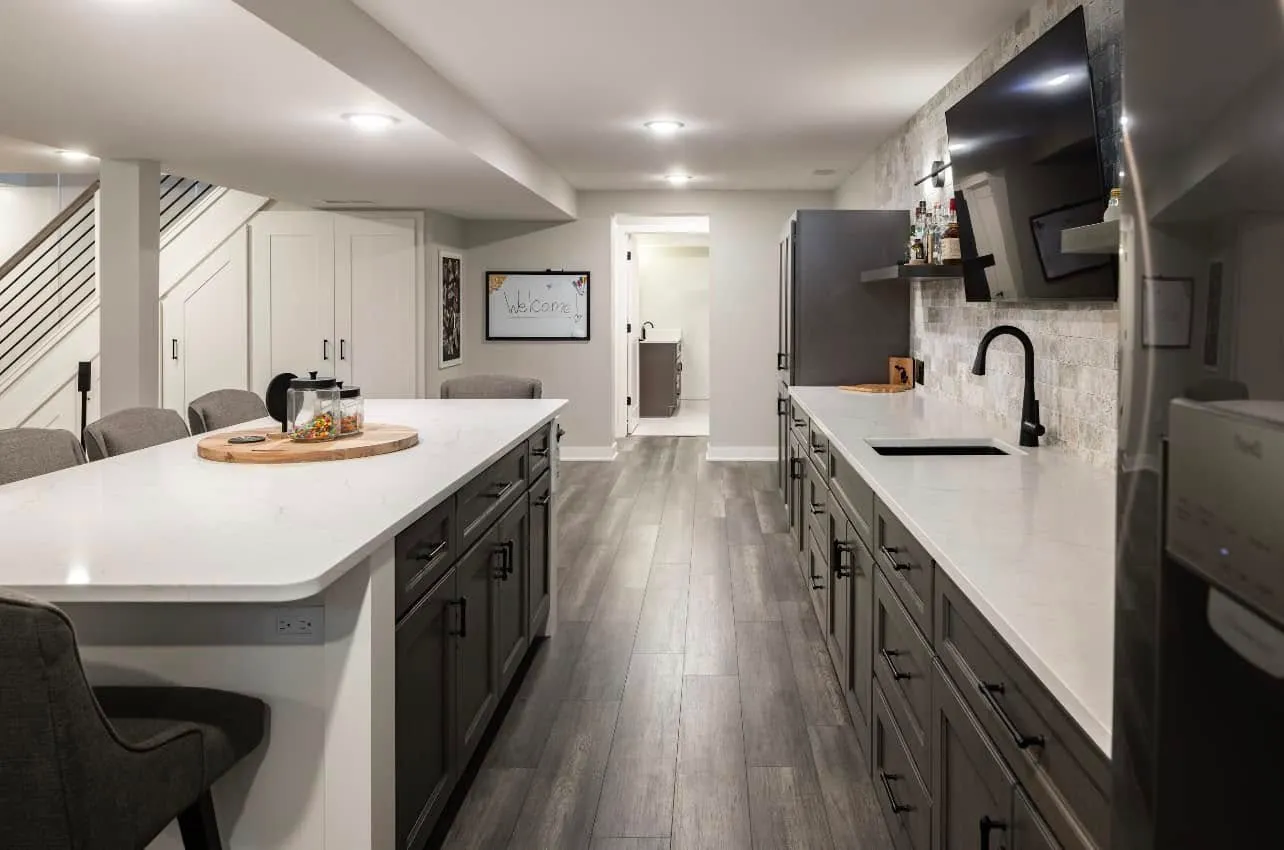 Finished basement bar with a white quartz island, dark cabinetry, black faucet, stone backsplash, wall-mounted TV, and gray wood-look flooring under the stairs.
