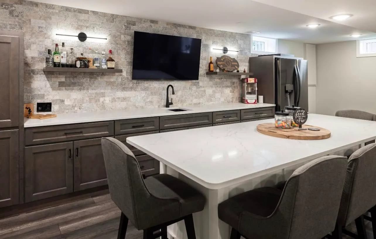 Basement kitchenette with gray cabinets, quartz counters, stone-tile backsplash, floating liquor shelves, mounted TV, and a white quartz island with gray stools.