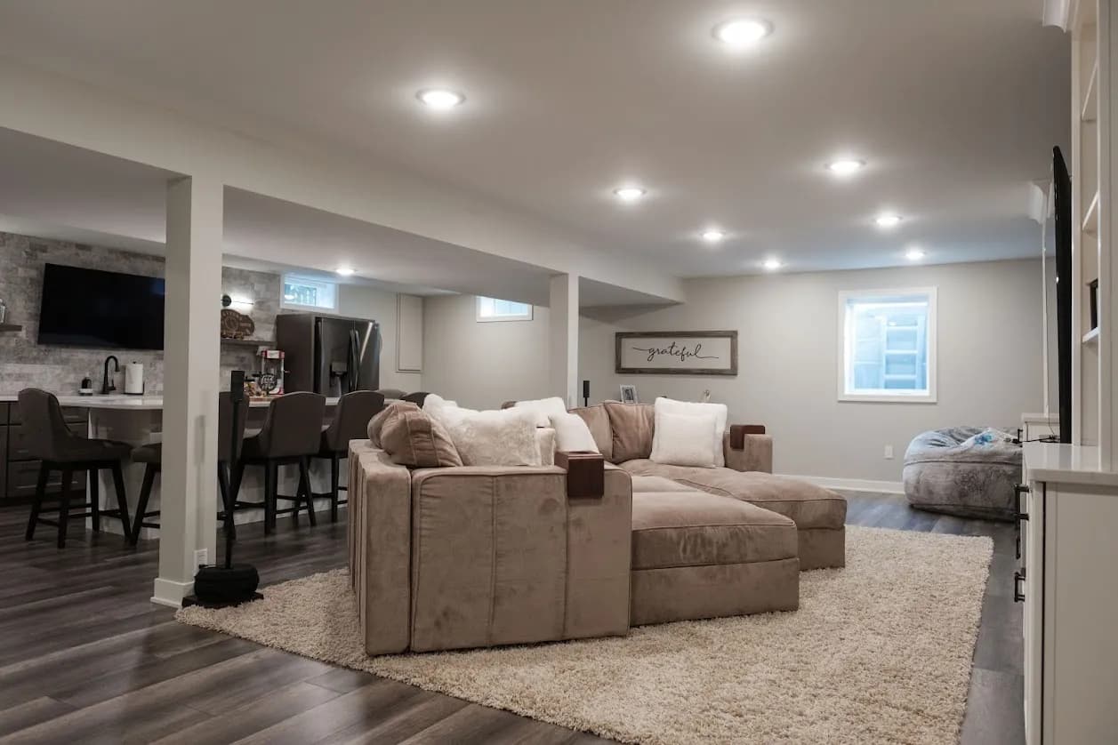 Open basement lounge with a large tan sectional on a shag rug, recessed lighting, a gray beanbag, and a bar area with stools in the background.