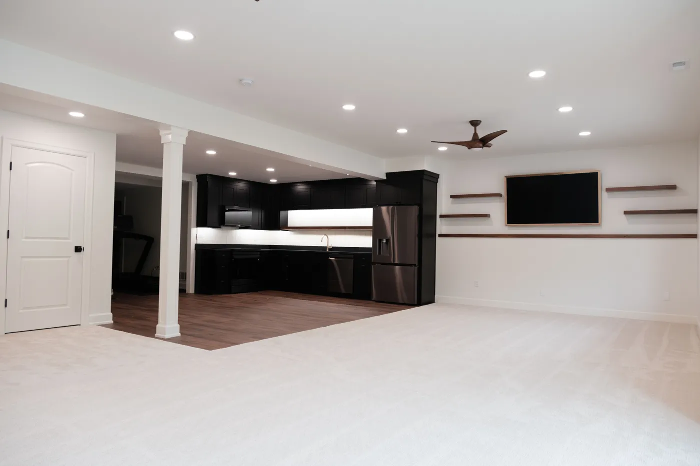 Newly finished basement with cream carpet, a black L-shaped kitchenette, stainless refrigerator, ceiling fan, floating shelves, and a framed mounted TV.