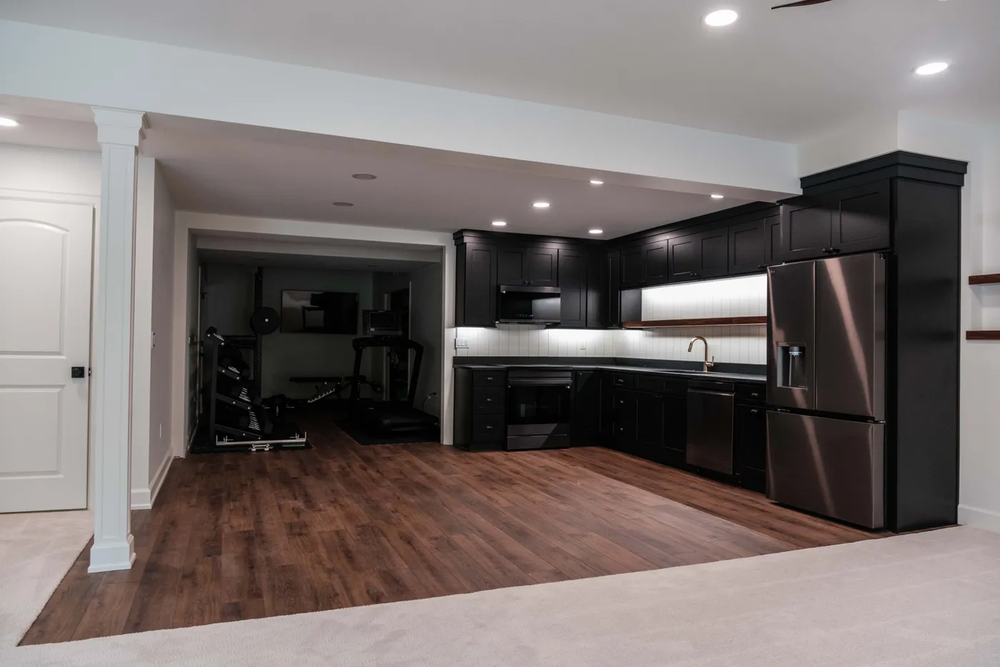 Basement kitchenette in black cabinetry with stainless appliances, tile backsplash, wood-look flooring, and an adjoining home gym with a treadmill and weights.