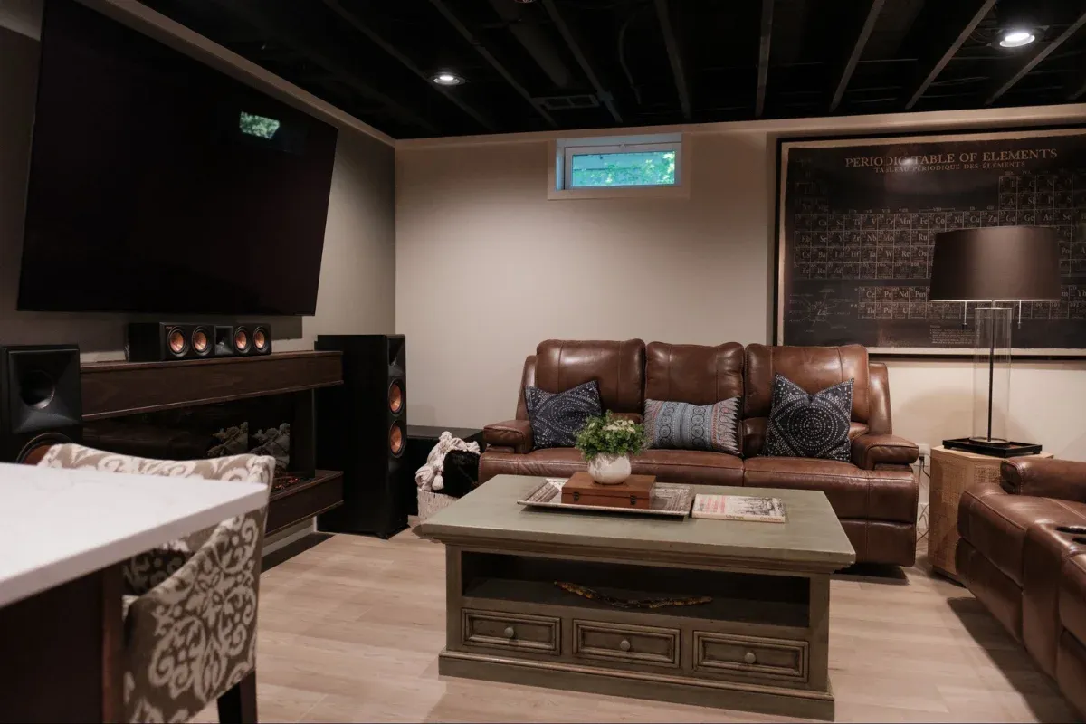 Cozy basement media room with brown leather sofas, tower speakers, a large mounted TV, a green coffee table, and a periodic-table poster on the ceiling.