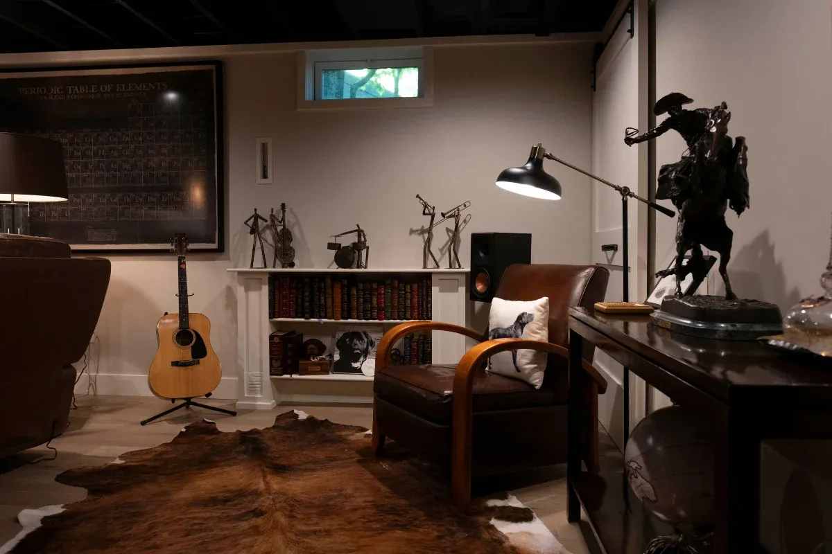 Basement den corner with a leather club chair, built-in bookshelves, an acoustic guitar, a cowhide rug, and a bronze horse sculpture.