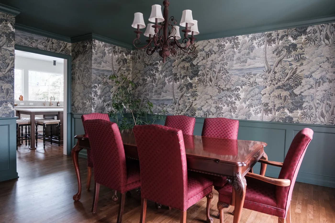 Formal dining room with botanical toile wallpaper above deep teal wainscoting, red upholstered chairs around a mahogany table beneath an iron chandelier.