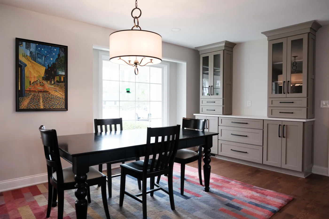 Dining area with paired gray glass-front hutch cabinets and drawers flanking a window, and a black table and chairs on a striped rug.