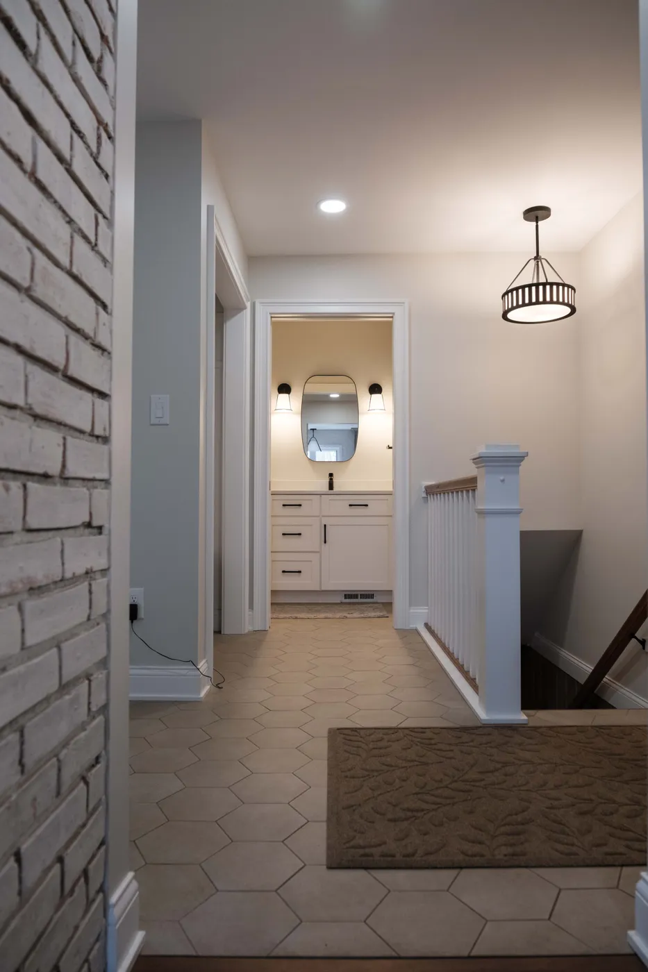 Upstairs hallway with hexagon tile floor, a whitewashed brick wall, and a lantern pendant leading to a vanity with a framed mirror and stair railing.