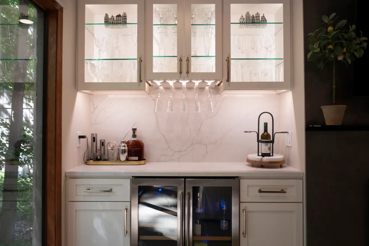 Built-in wet bar with glass-front upper cabinets, a marble slab backsplash, hanging stemware, dual glass-door beverage fridges, and countertop bar tools.