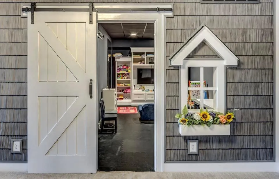 Basement children's playhouse built with gray shingle siding, a sliding barn door, and a peaked faux window with a sunflower flower box.