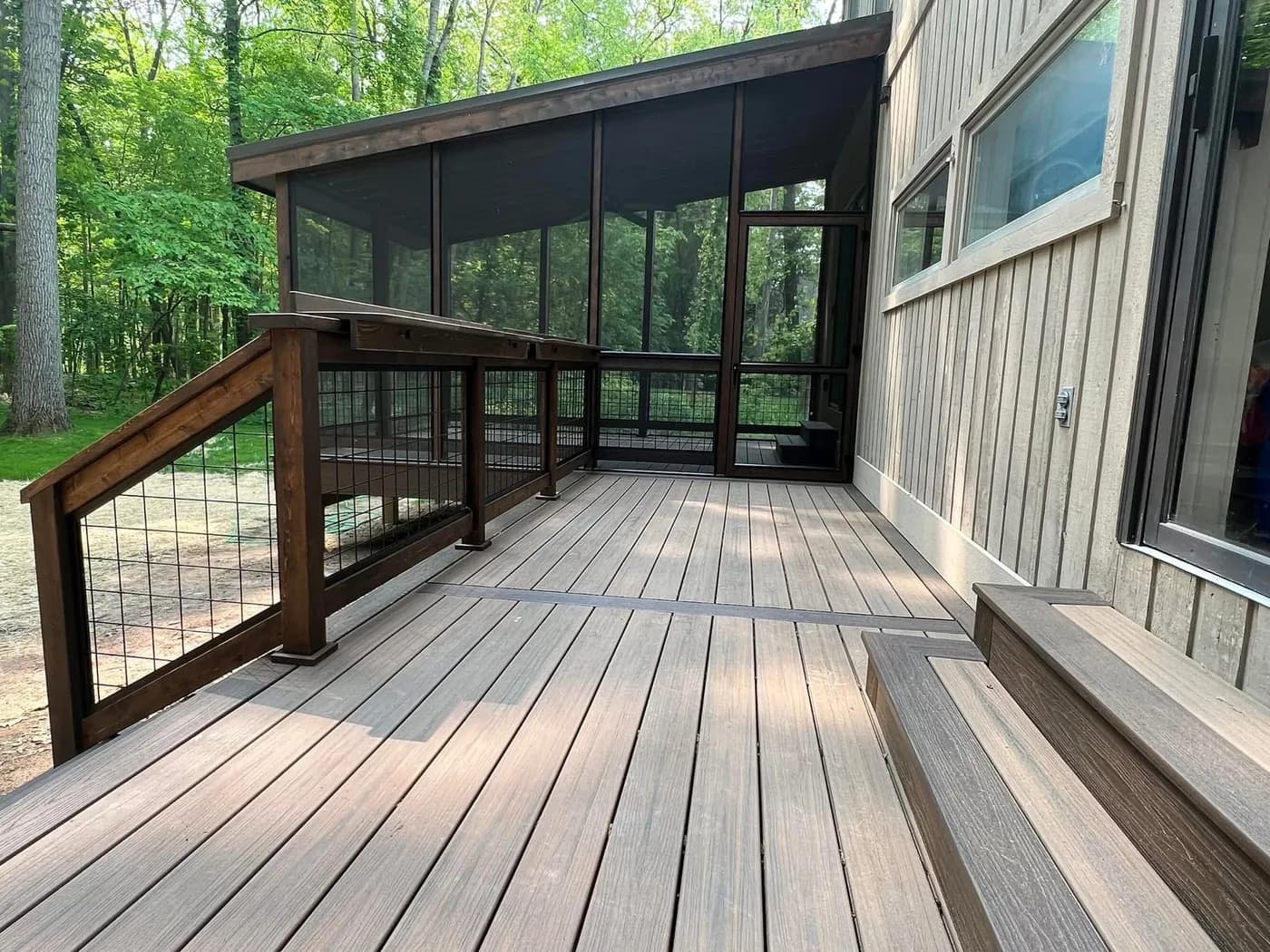 Composite wood-grain deck with dark-stained wood railings and hog-wire panels beside a screened porch, backing onto green forest.