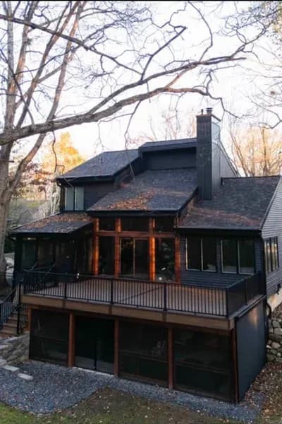 Rear exterior of a dark-sided home showing a raised deck above a screened lower porch, framed by bare autumn trees at dusk.