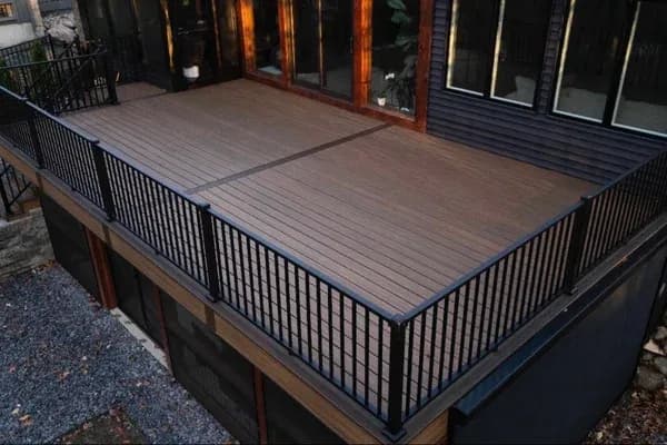 Elevated composite deck with brown wood-grain boards and black metal balusters wrapping the corner of a dark home over a stone base.