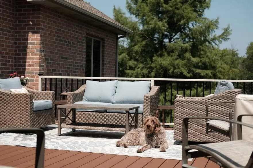 Sunny brick-home deck with woven wicker furniture, blue cushions, a patterned outdoor rug, and a dog lounging in the center.