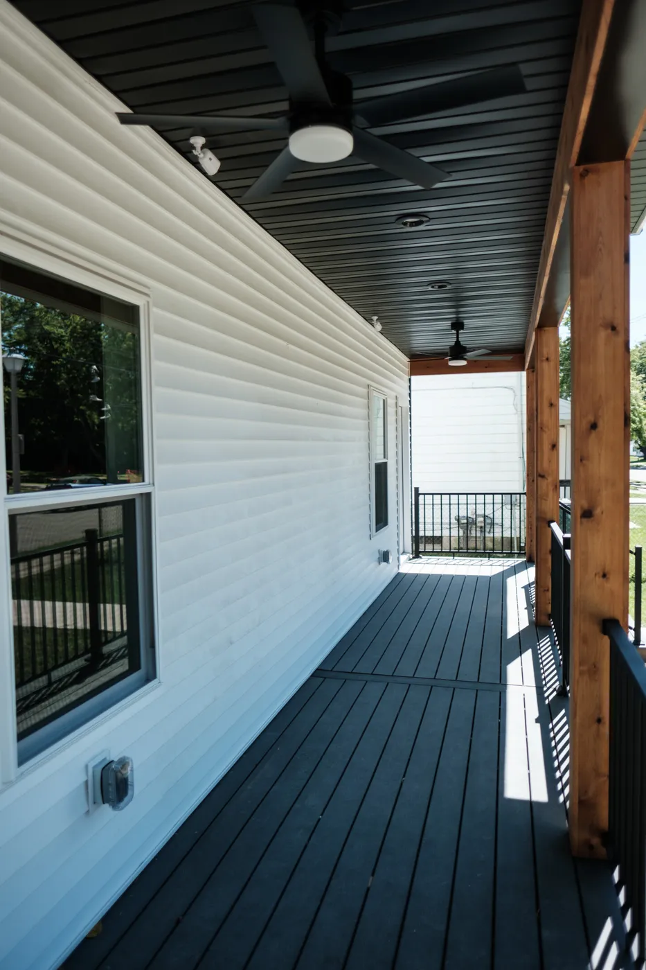 Covered front porch with dark composite flooring, white siding, natural cedar posts, a black metal railing, and a black ceiling fan overhead.