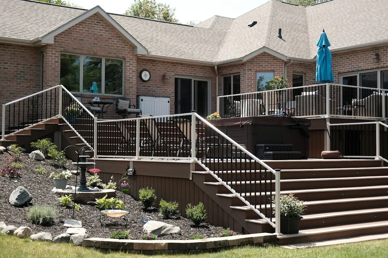 Two-tier composite deck with white railings, a hot tub, and landscaped beds behind a brick home
