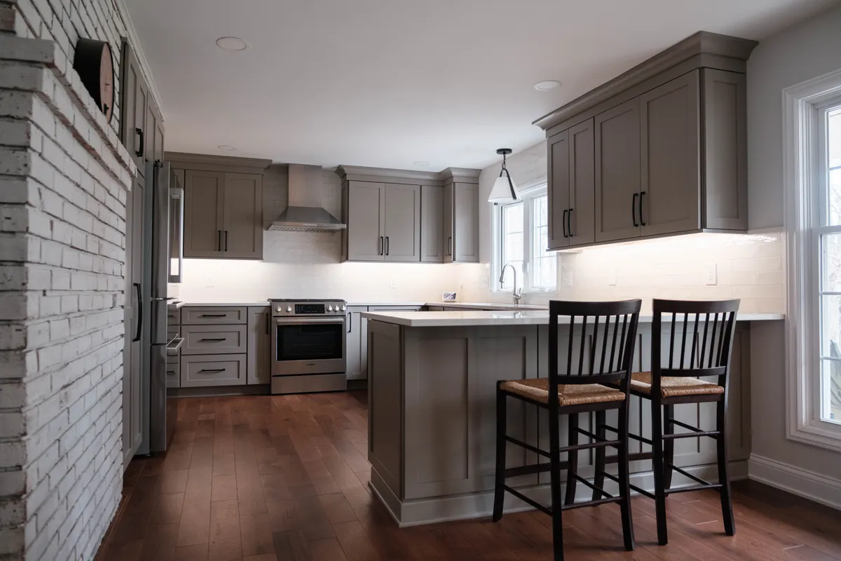 Wide view of the finished remodel — grey shaker cabinetry, quartz peninsula and the whitewashed brick wall